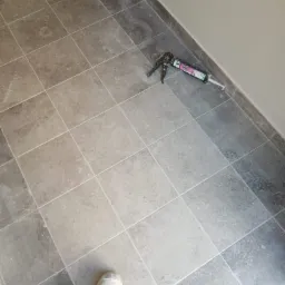 Ułożona podłoga z szarych płytek ceramicznych, pistolet do silikonu leżący przy ścianie, widoczny fragment buta robotnika.