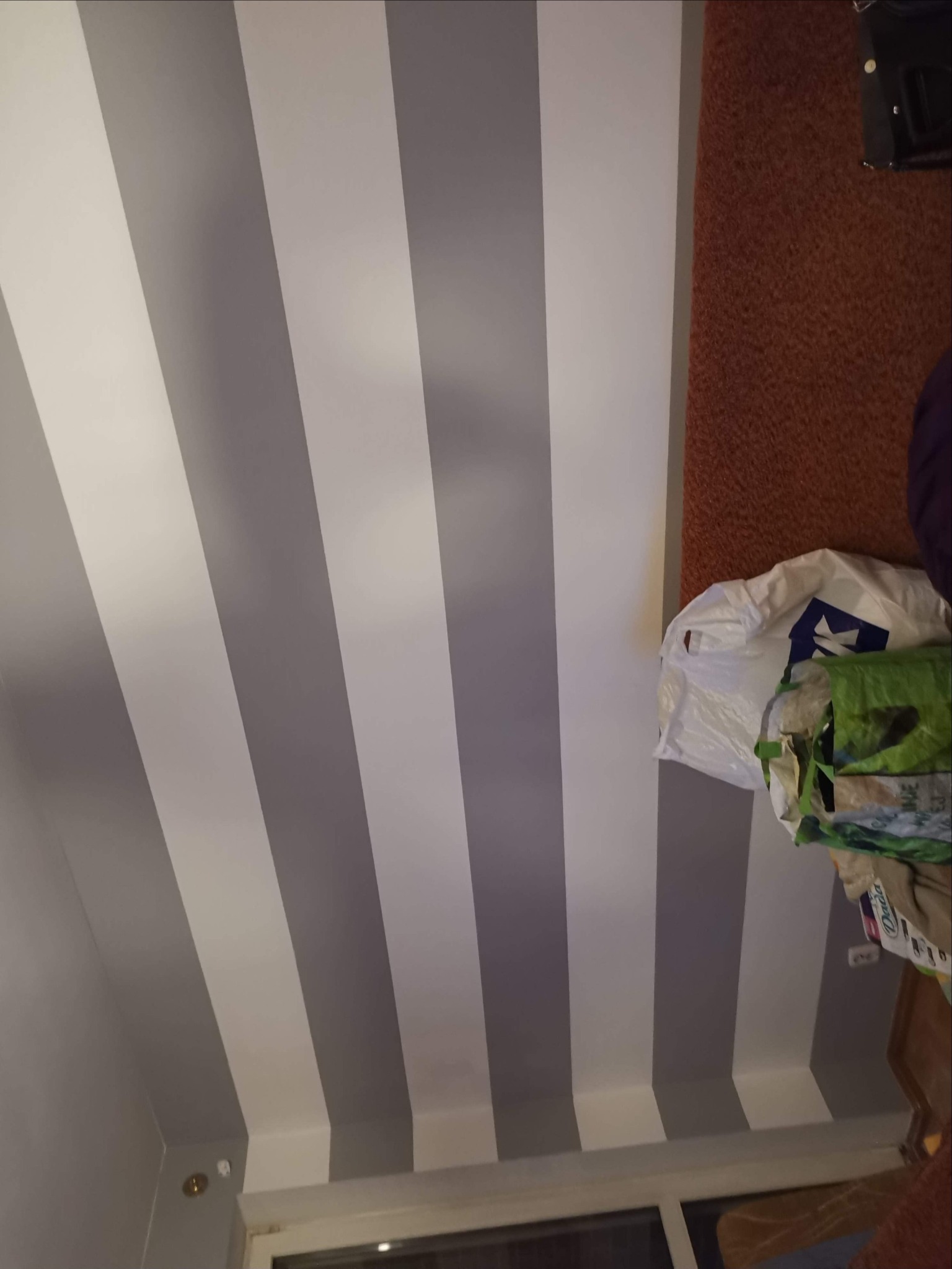 Ściana pomalowana w szerokie, pionowe pasy na szaro i biało, widoczne worki z farbą i narzędzia malarskie, sugerujące świeżo zakończone prace.