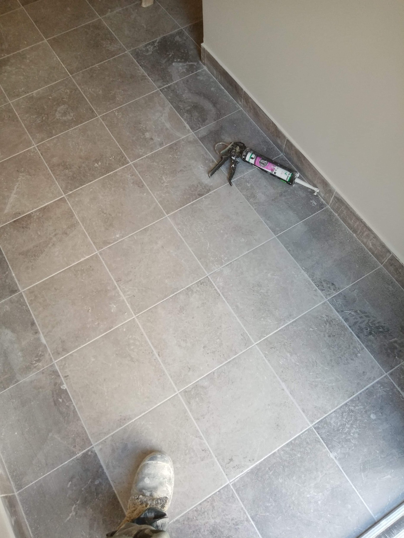 Ułożona podłoga z szarych płytek ceramicznych, pistolet do silikonu leżący przy ścianie, widoczny fragment buta robotnika.