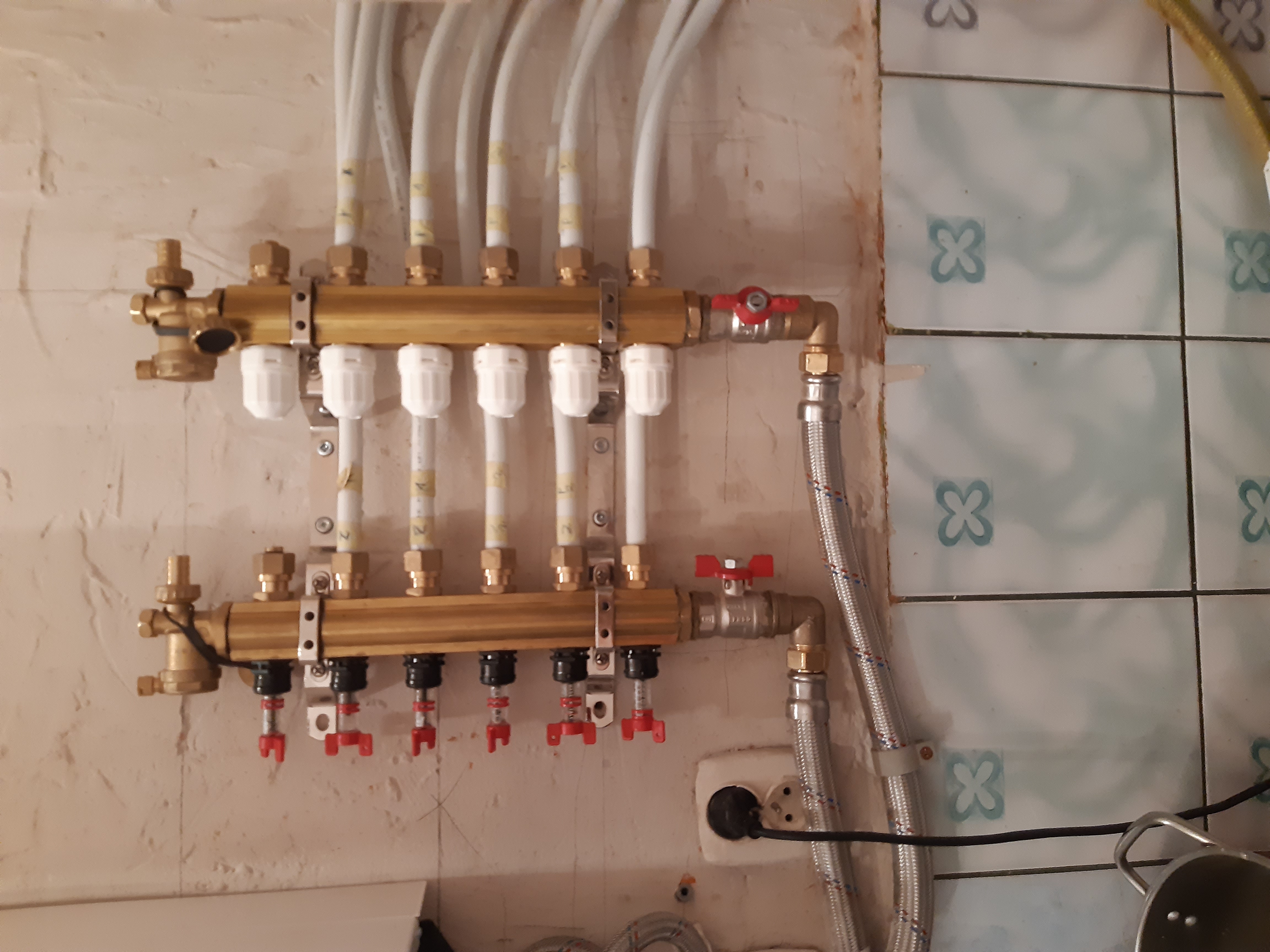 Rozdzielacz centralnego ogrzewania zamontowany na ścianie, widoczne rury doprowadzające wodę i zawory regulacyjne, obok fragment ściany z płytkami w kwiatki.