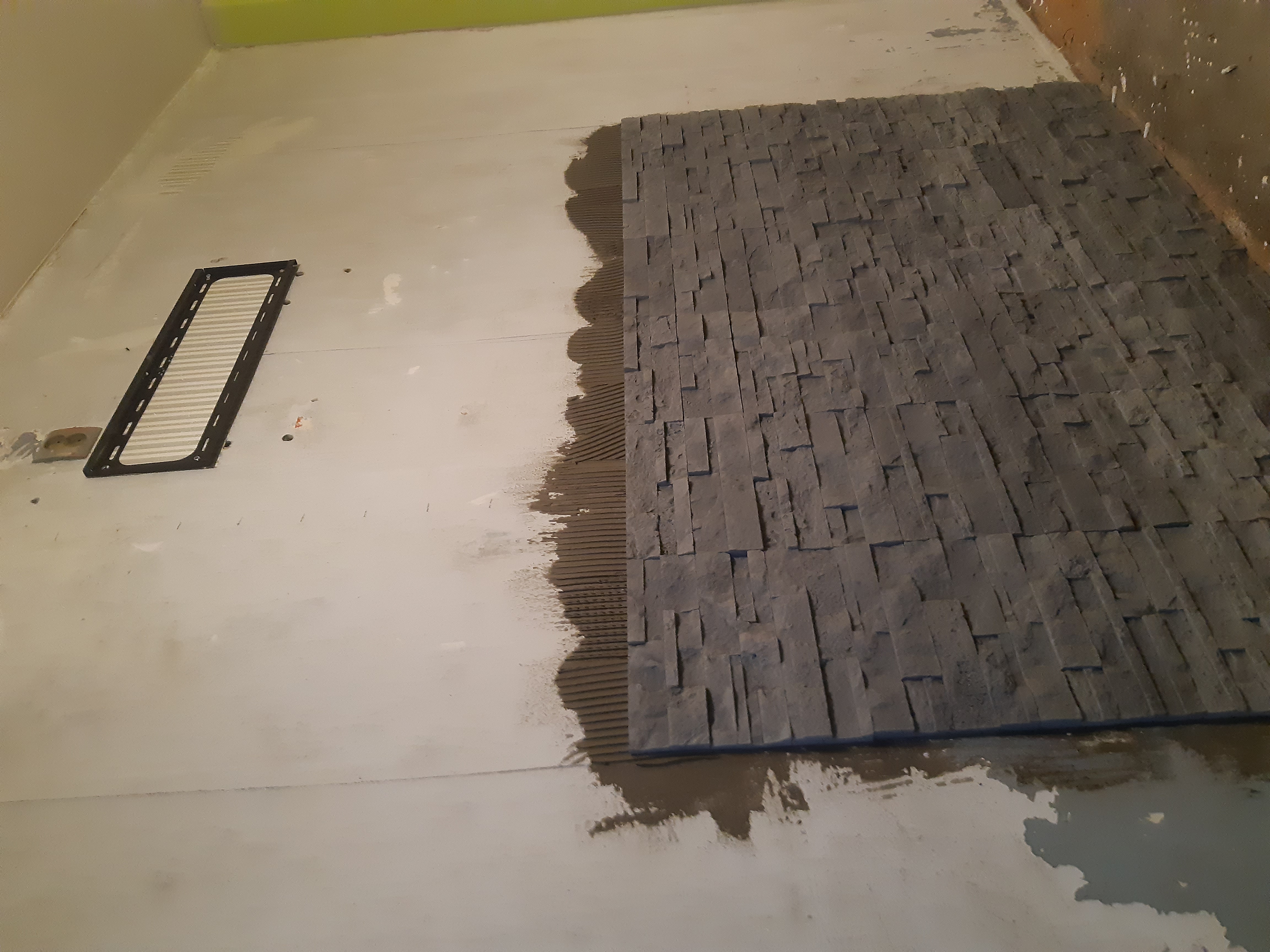 Montaż dekoracyjnej okładziny kamiennej na ścianie wewnętrznej, widoczny klej i fragment ułożonej powierzchni z nieregularnych, szarych elementów.