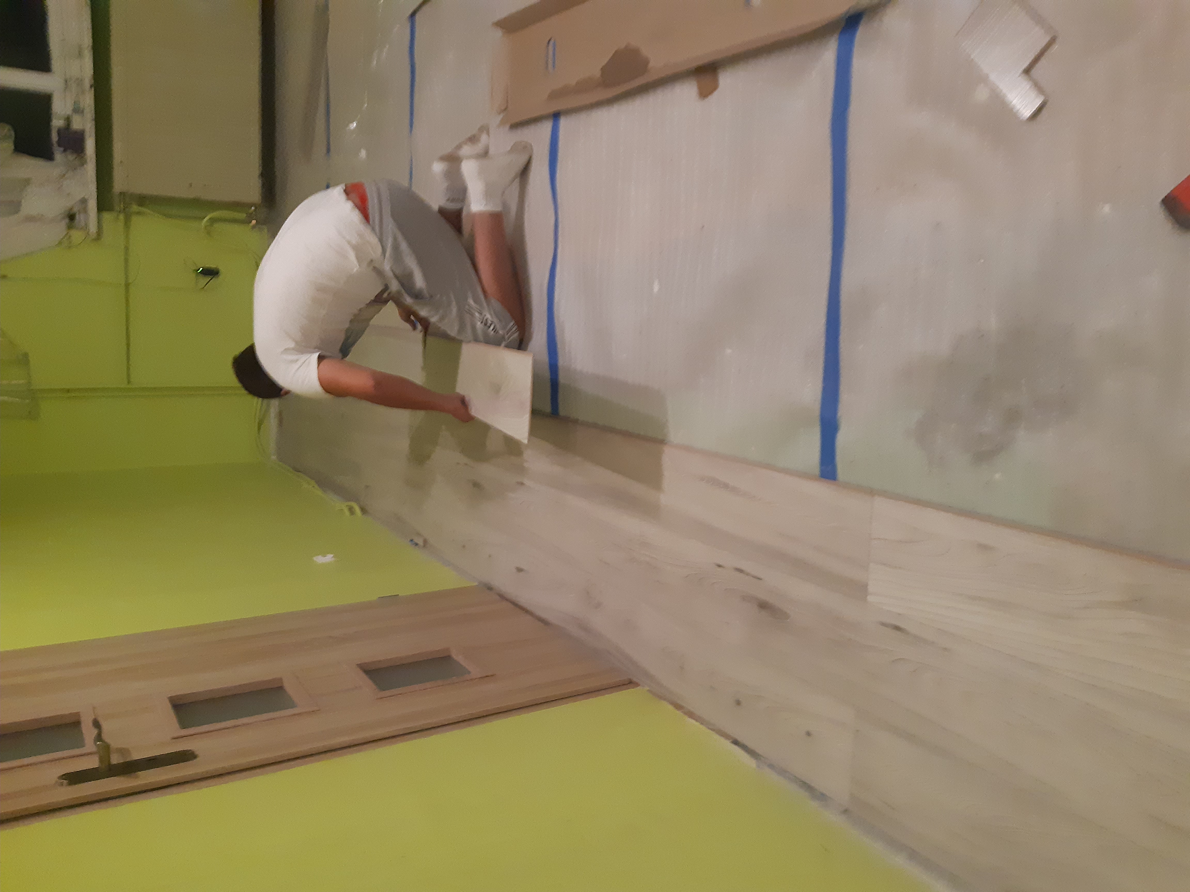 Pracownik w trakcie układania paneli podłogowych na jaskrawozielonej posadzce, widoczne panele z otworami na drzwi i niebieska taśma malarska na ścianie.