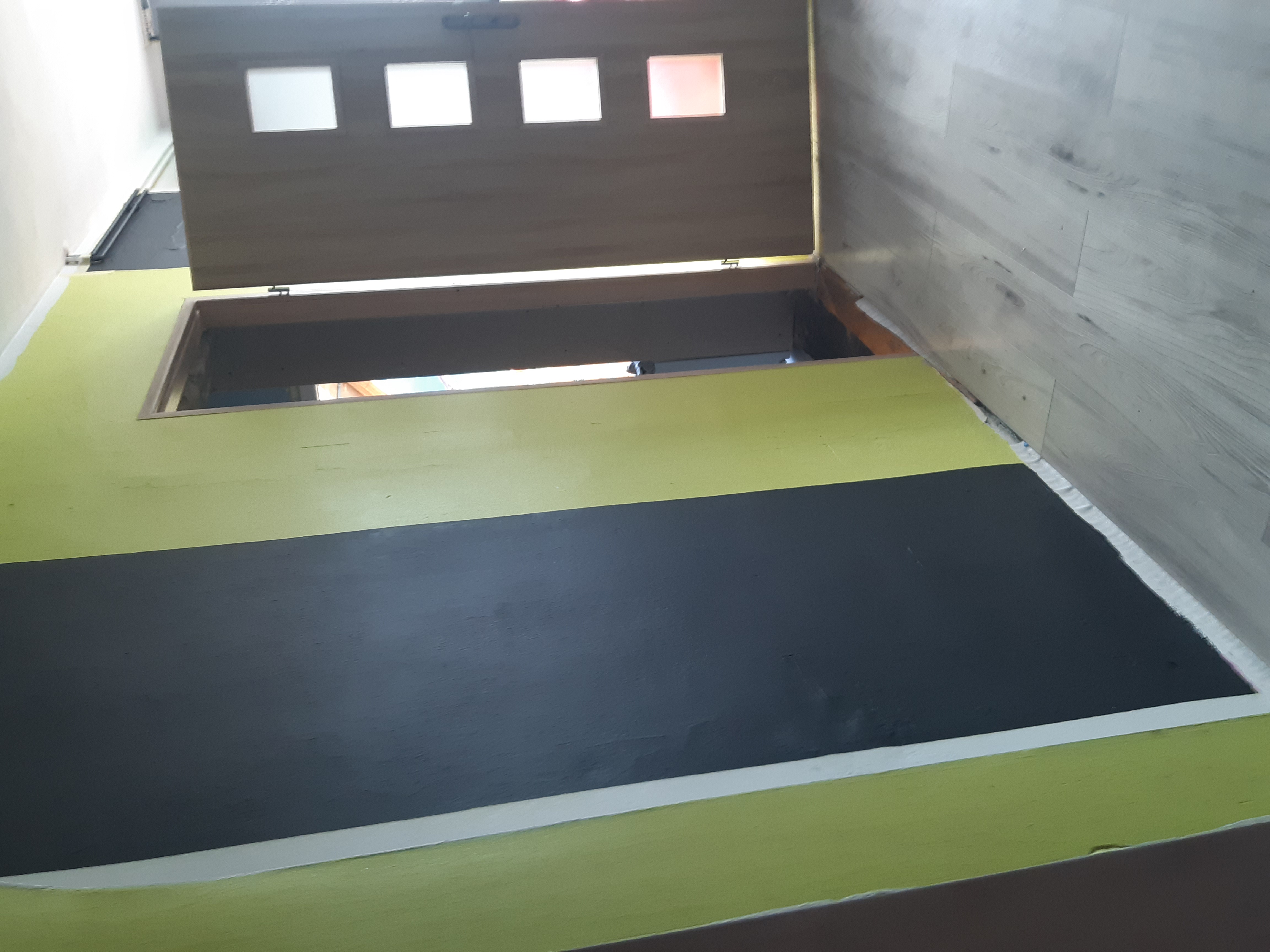 Otwarty właz do piwnicy z drewnianą klapą z szybkami, pomalowany fragment podłogi na zielono i czarno, ściana wykończona panelami podłogowymi.