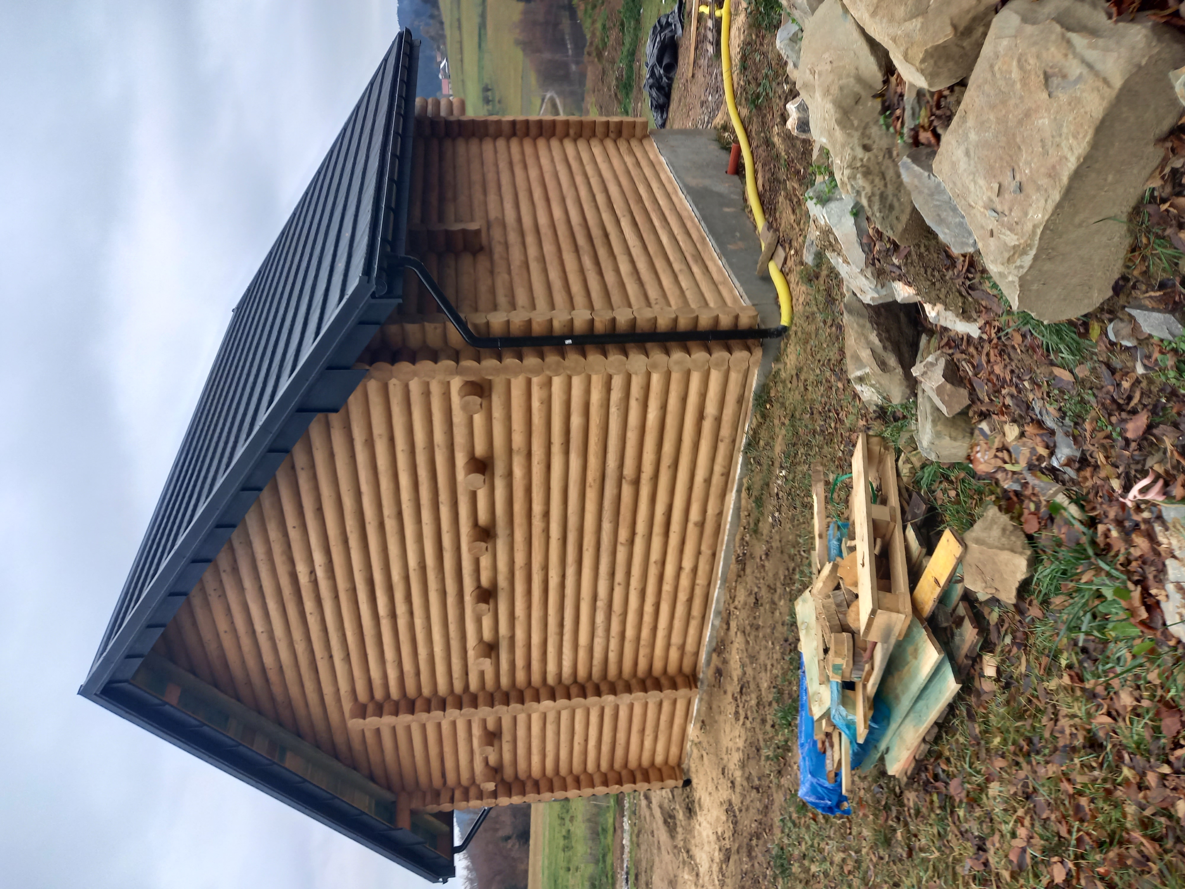 Gotowa drewniana altana z bali z czarnym dachem i rynną, widok z boku na tle zieleni i pochmurnego nieba, obok leżą skały i palety.