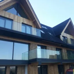 Nowoczesny budynek z drewnianą elewacją i balkonami zabezpieczonymi szklanymi balustradami, widok z dołu na elewację budynku z oknami i balkonami.