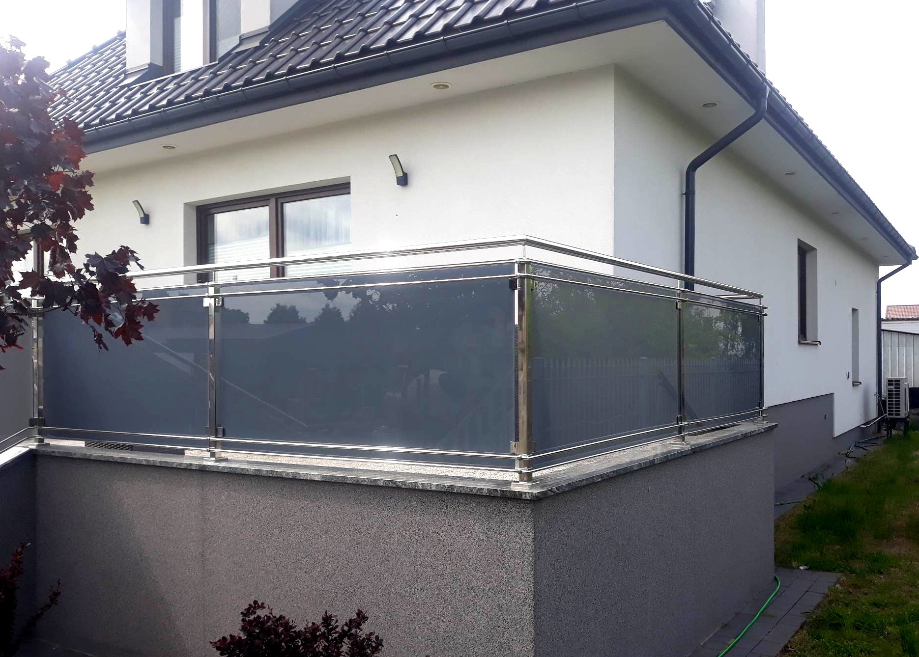 Balkon z nowoczesną balustradą ze stali nierdzewnej i szarym szkłem, widok z zewnątrz na tle białego domu z ciemną dachówką.