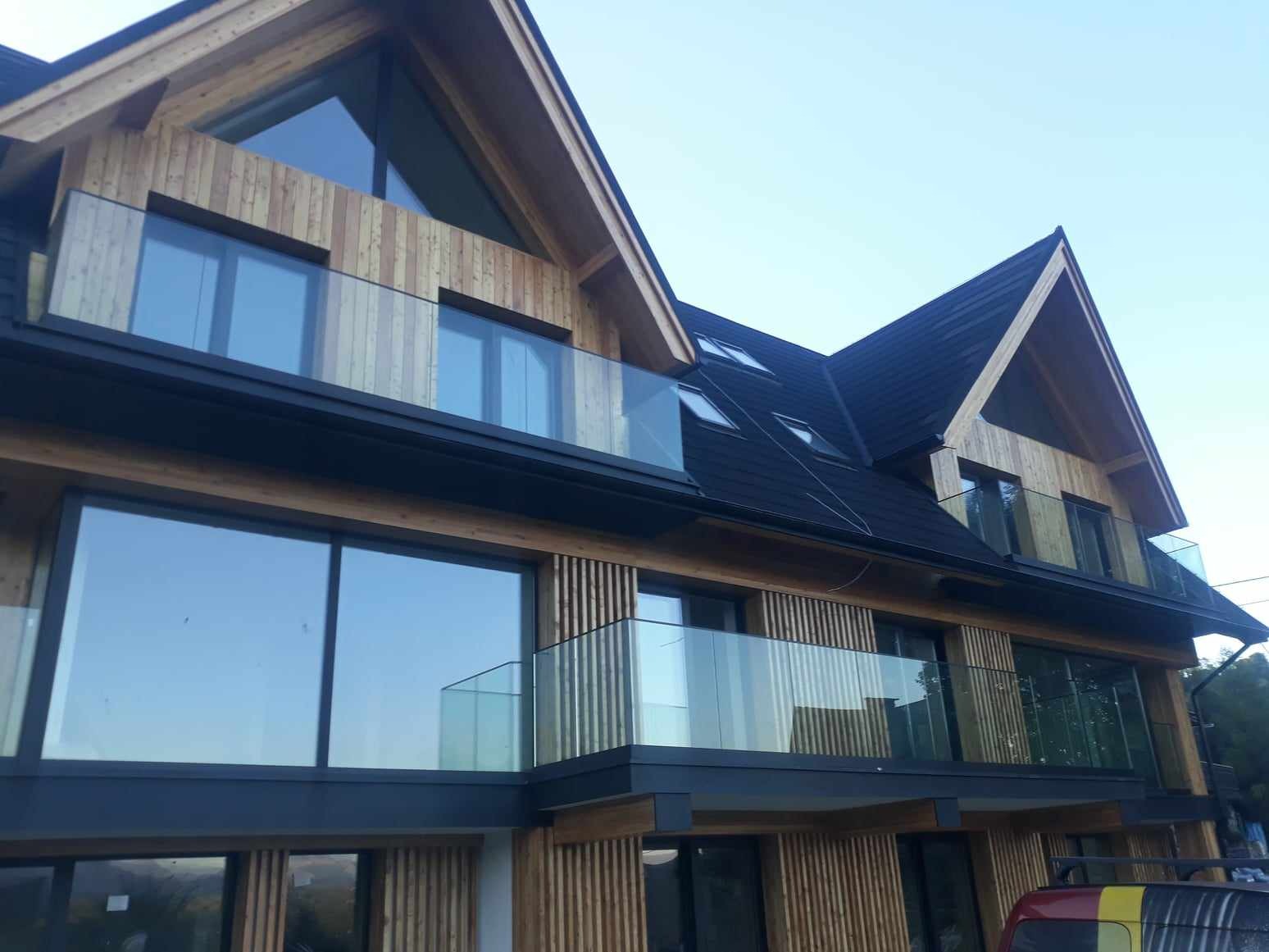 Nowoczesny budynek z drewnianą elewacją i balkonami zabezpieczonymi szklanymi balustradami, widok z dołu na elewację budynku z oknami i balkonami.