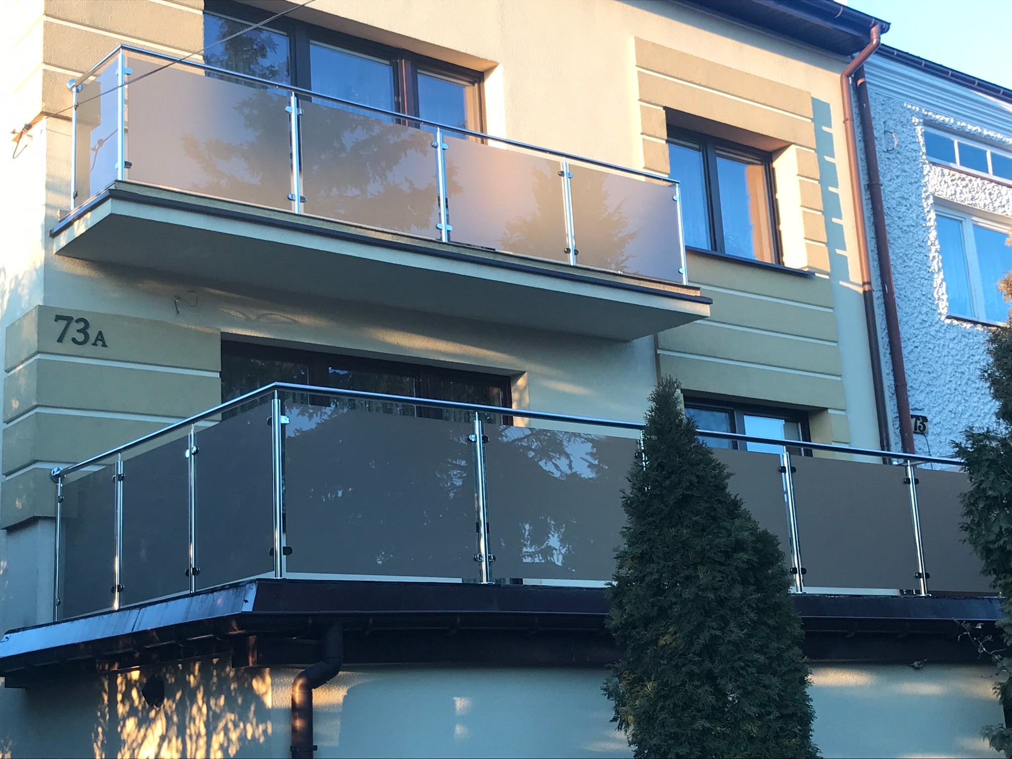 Elewacja budynku z dwoma balkonami zabezpieczonymi szklanymi panelami w stalowych ramach, widoczne numery adresowe 73A i 75.