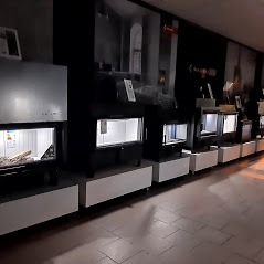 Rząd nowoczesnych wkładów kominkowych w salonie ekspozycyjnym, oświetlonych od wewnątrz, z widocznymi elementami imitującymi drewno. W tle ciemne ściany z grafiką i oznaczeniami.