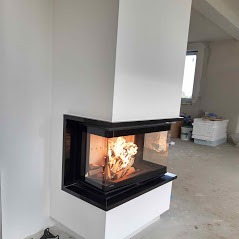 Narożny wkład kominkowy z czarną obudową i widocznym płomieniem, umieszczony w surowym, białym wnętrzu przed wykończeniem.