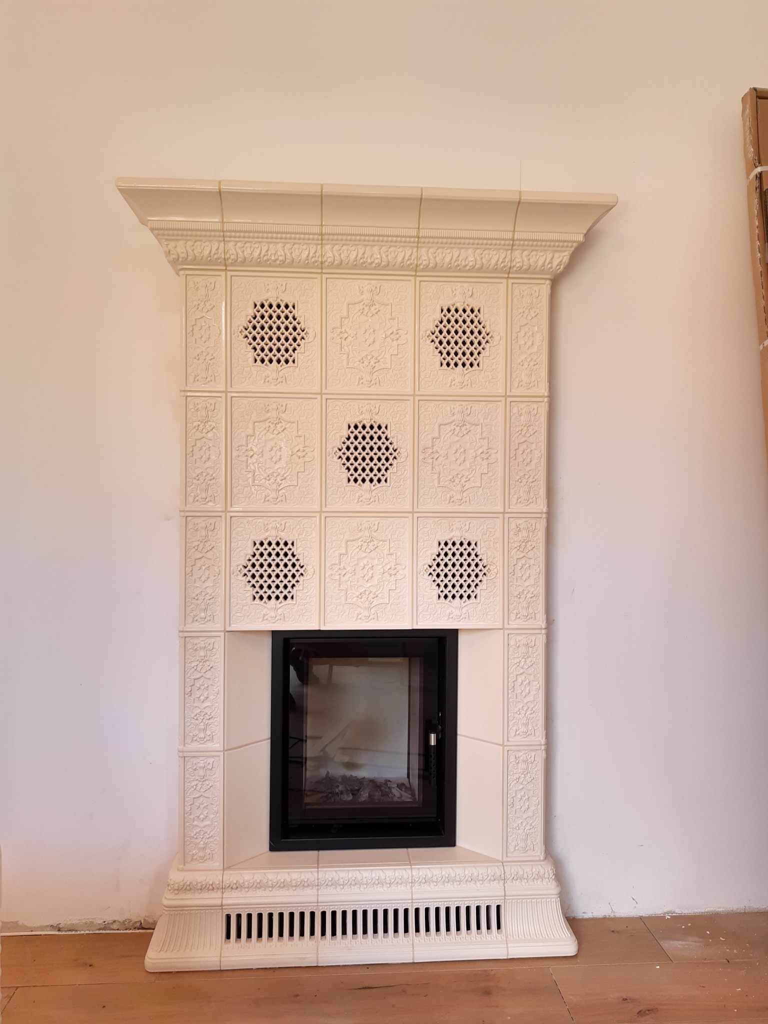 Kremowy, kaflowy kominek z ozdobnymi ażurowymi panelami i czarnym wkładem kominkowym, zamontowany w Białymstoku, na tle białej ściany i drewnianej podłogi.