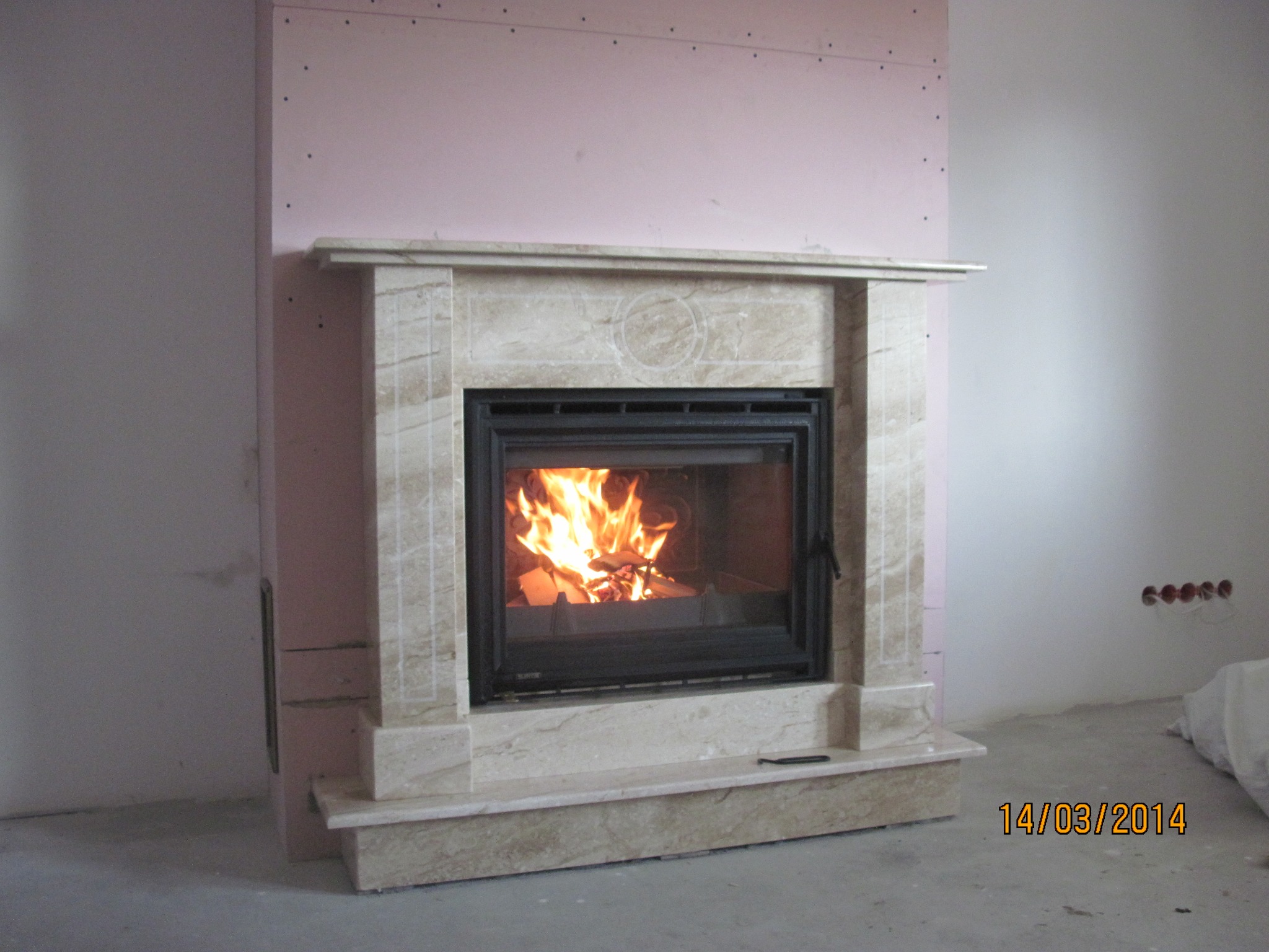 Wnętrze z marmurową obudową w trakcie montażu, z paleniskiem z płomieniami, widoczne płyty gipsowe na ścianie i niedokończone wykończenie.