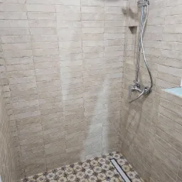 Kabina prysznicowa wyłożona beżowymi, podłużnymi płytkami na ścianach i wzorzystymi, niebiesko-białymi płytkami na podłodze z odpływem liniowym, bateria prysznicowa z deszczownicą i półka w ścianie.