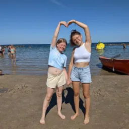 Dwie nastolatki trzymają się za ręce na plaży, tworząc serce z rąk nad głowami, w tle morze, pomarańczowa łódź ratunkowa i osoby w wodzie.