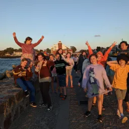 Grupa roześmianych dzieci i dorosły pozują na tle morza, falochronu i budynku z wieżą podczas zachodu słońca.