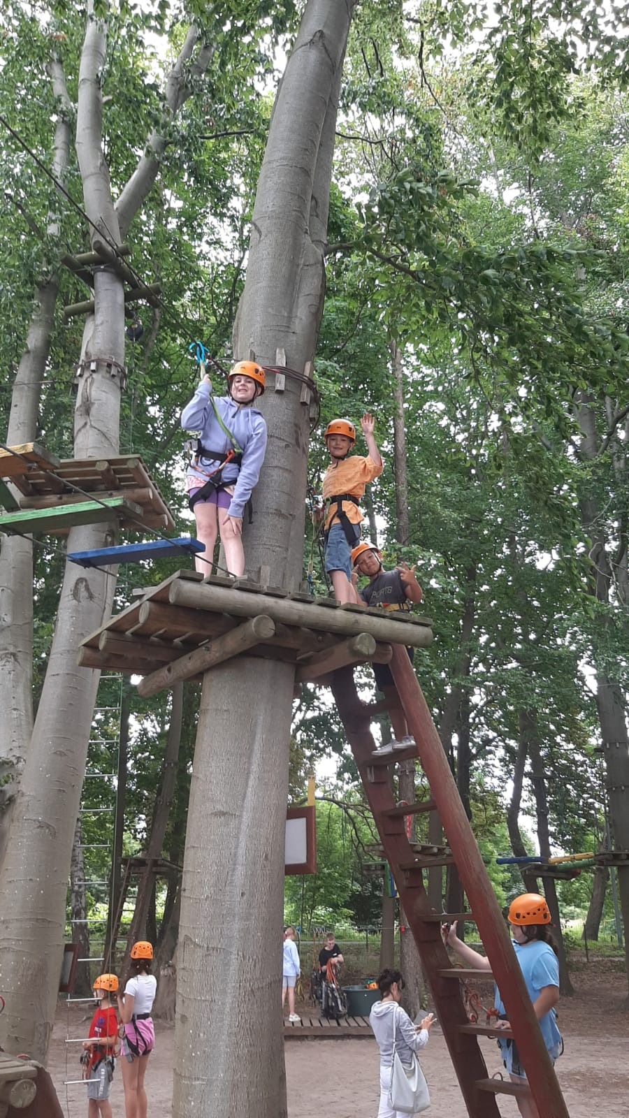 Grupa dzieci w kaskach i uprzężach na platformach w parku linowym, jedno dziecko macha, a inne pokazuje znak pokoju na tle drzew.