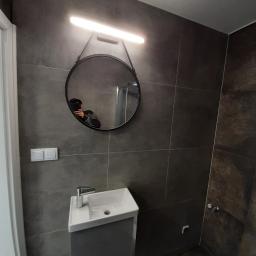 AQUA-TERM2 - Nowoczesna łazienka z ciemnymi, płytkami imitującymi kamień, okrągłym lustrem z podświetleniem LED, białą umywalką nablatową na szarej szafce i widocznym odbiciem osoby robiącej zdjęcie.