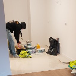 AQUA-TERM2 - Pracownik w bluzie z kapturem sięga do skrzynki narzędziowej w trakcie prac w pomieszczeniu z białymi ścianami, widoczne puszki z farbą, czarny worek na śmieci, żółta kamizelka odblaskowa i płytki...