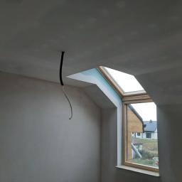 AQUA-TERM2 - Poddasze w trakcie zabudowy: surowe ściany z widocznymi przewodami elektrycznymi, okno dachowe z widokiem na dom sąsiada.