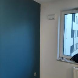 AQUA-TERM2 - Pomieszczenie po malowaniu ścian na biało i granatowo, widoczny grzejnik, okno z widokiem na budynek oraz gniazdko elektryczne na granatowej ścianie.