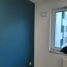 Pomieszczenie po malowaniu ścian na biało i granatowo, widoczny grzejnik, okno z widokiem na budynek oraz gniazdko elektryczne na granatowej ścianie.