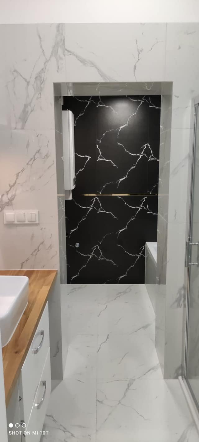 Nowoczesna łazienka we Wrocławiu z białymi marmurowymi płytkami na ścianach i podłodze, kontrastująca czarna ściana w kabinie prysznicowej, drewniany blat pod umywalką i białe szafki.