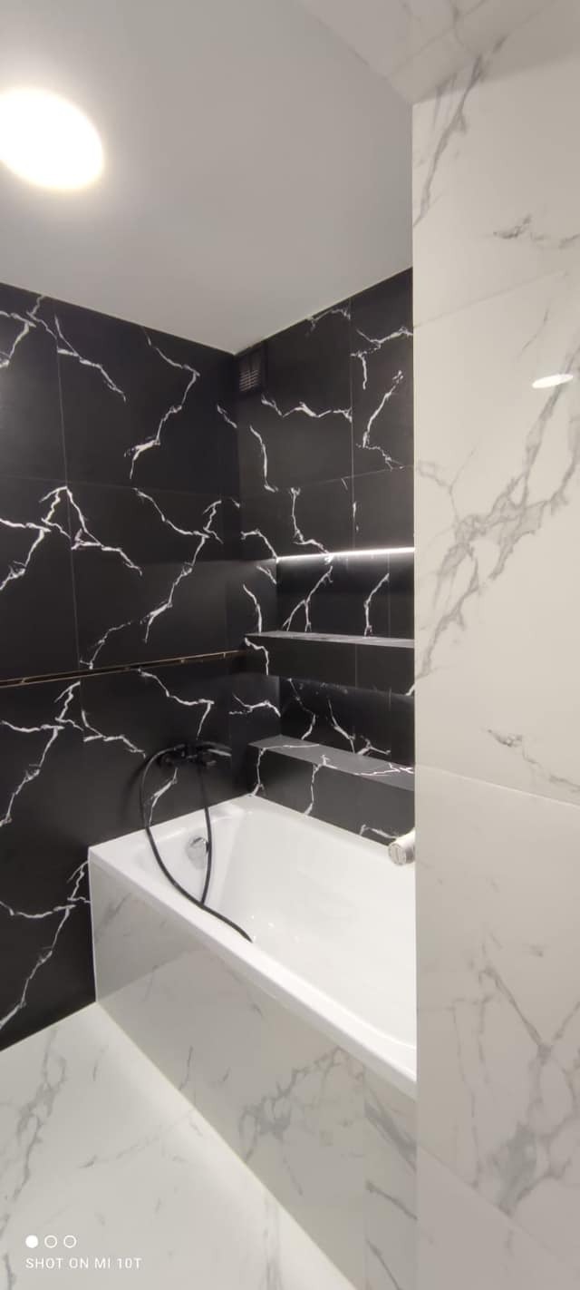 Nowoczesna łazienka z wanną obłożoną białymi płytkami imitującymi marmur, czarne płytki z białymi żyłami na ścianach i półkach, oraz czarna bateria wannowa.