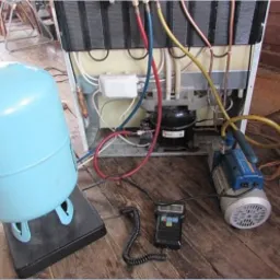 Tył lodówki z widocznym agregatem, podłączony do butli z czynnikiem chłodniczym i pompy próżniowej, miernik elektroniczny leży obok.