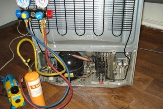 Tył lodówki podczas serwisu: podłączone manometry i butla z czynnikiem chłodniczym R600a, widoczny kompresor i skraplacz.