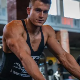 Mężczyzna w czarnej koszulce bez rękawów pozuje w siłowni, opierając się o sztangę, z widocznymi żyłami na ramionach i zarysowanymi mięśniami.