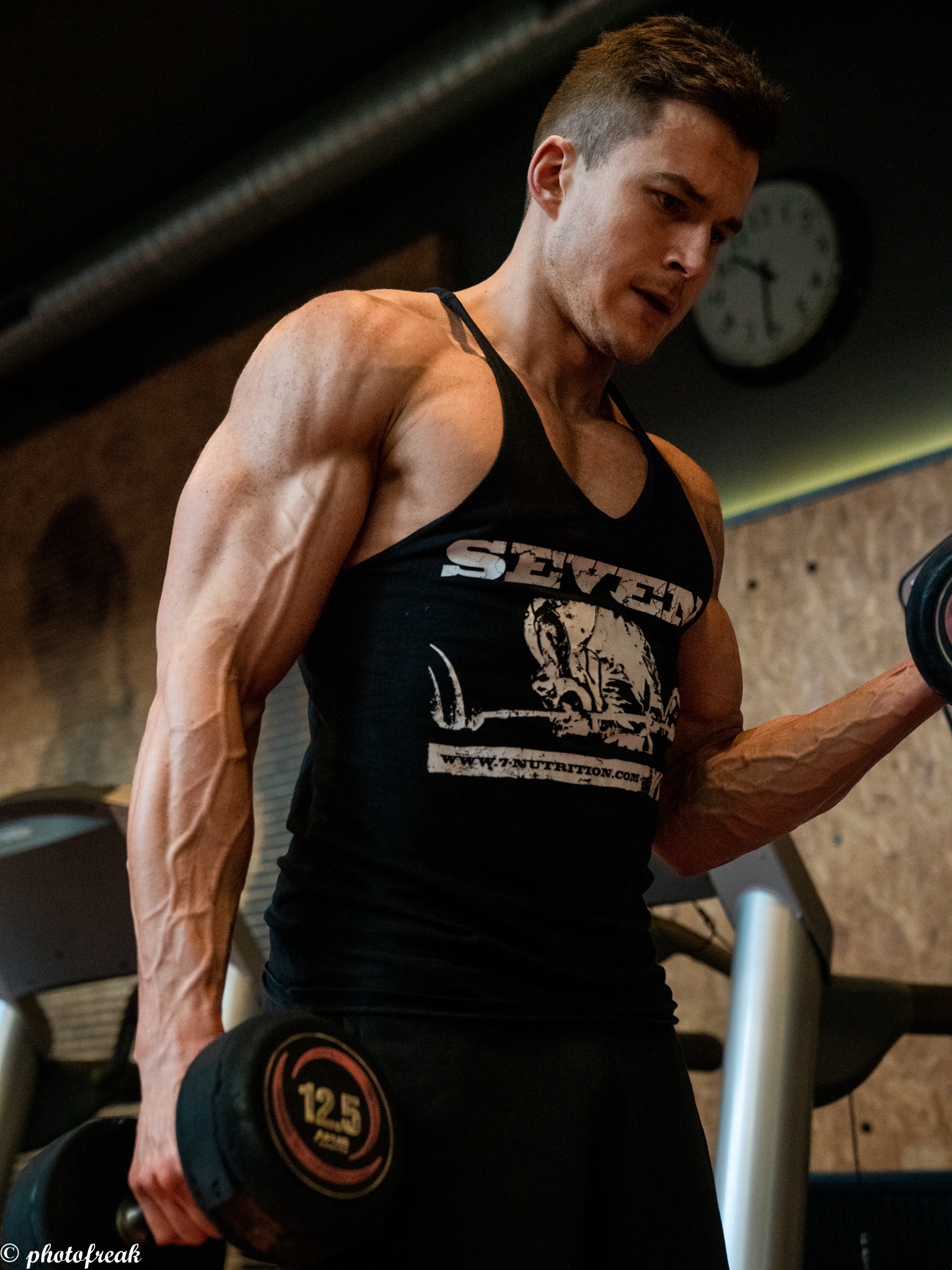 Mężczyzna z wyraźnie zarysowanymi mięśniami, w czarnym tank topie z logo, ćwiczy z hantlą 12.5 kg w siłowni, widoczne bieżnie w tle i zegar na ścianie.