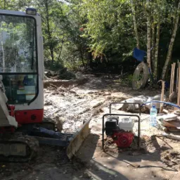 Mała koparka na gąsienicach stoi na błotnistym terenie, obok generatora i resztek szalunku, w tle widoczne drzewa.