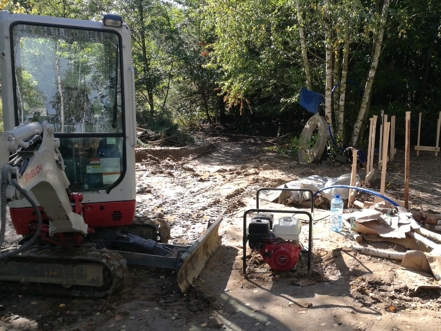 Mała koparka na gąsienicach stoi na błotnistym terenie, obok generatora i resztek szalunku, w tle widoczne drzewa.