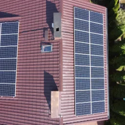 Widok z góry na instalację fotowoltaiczną na dachu z brązowej dachówki, obok komin i okno dachowe, widoczny fragment ogrodu.