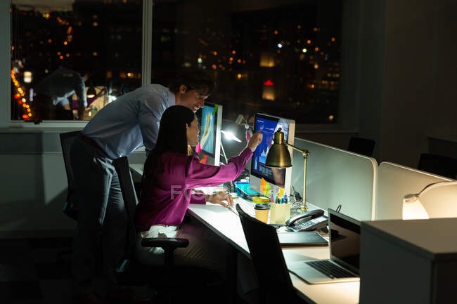 Dwoje pracowników biurowych analizuje dane na monitorze komputera w nowoczesnym biurze z widokiem na nocne miasto, przy oświetleniu lampki biurkowej.