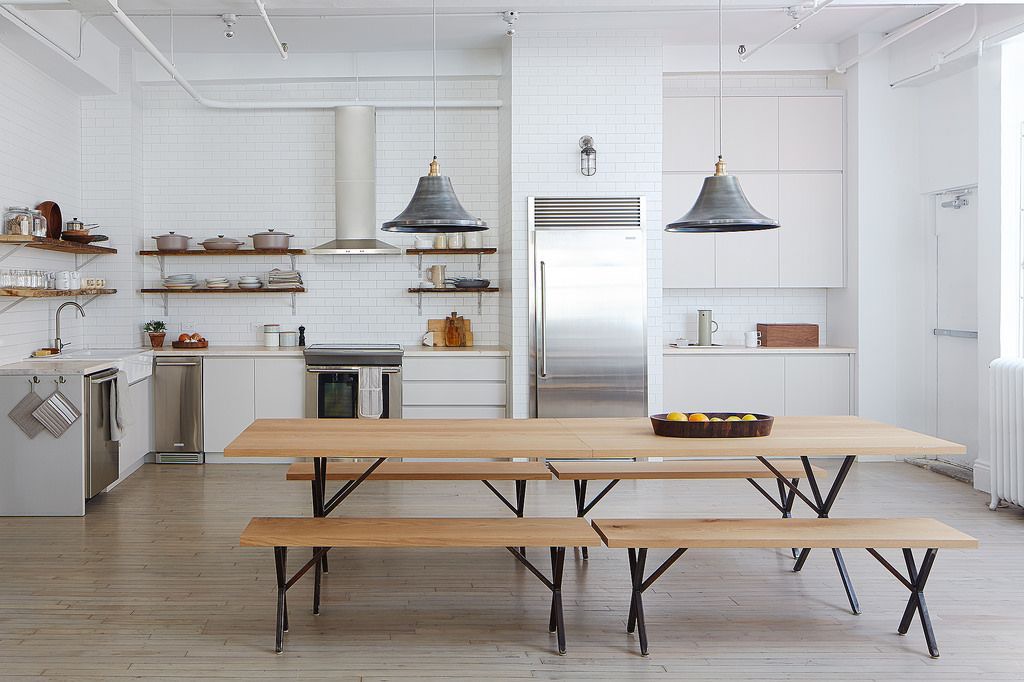 Jasna, przestronna kuchnia w Krakowie z białymi płytkami na ścianach, drewnianym stołem z ławkami na metalowych nogach, stalową lodówką i wiszącymi lampami w stylu industrialnym.