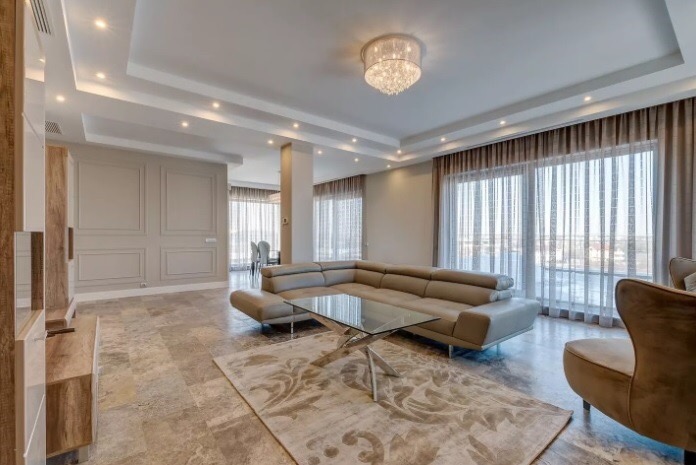 Elegancki salon z narożną sofą w odcieniach beżu, szklanym stolikiem kawowym na wzorzystym dywanie, marmurową podłogą i dużymi oknami z delikatnymi zasłonami.