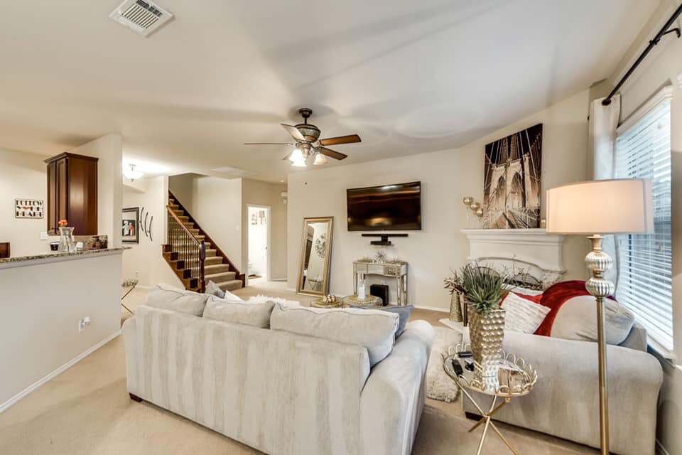 Jasny salon z beżowym dywanem, szarą kanapą, schodami z drewnianymi poręczami, telewizorem na ścianie i dekoracyjnym kominkiem z obrazem mostu Brooklińskiego