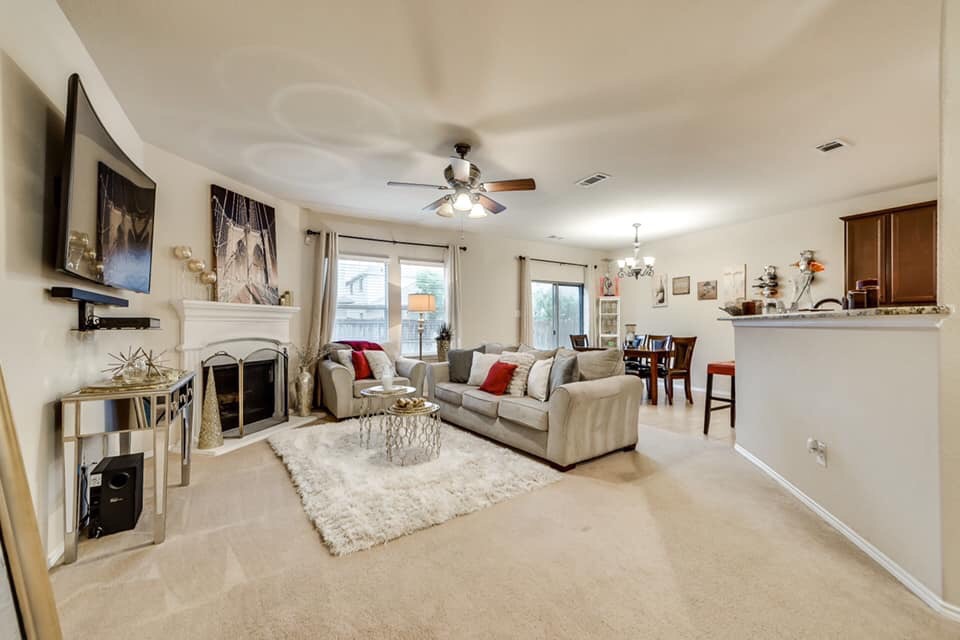 Jasne wnętrze salonu z beżową sofą, kominkiem i telewizorem na ścianie. W tle widoczna jadalnia i kuchnia.