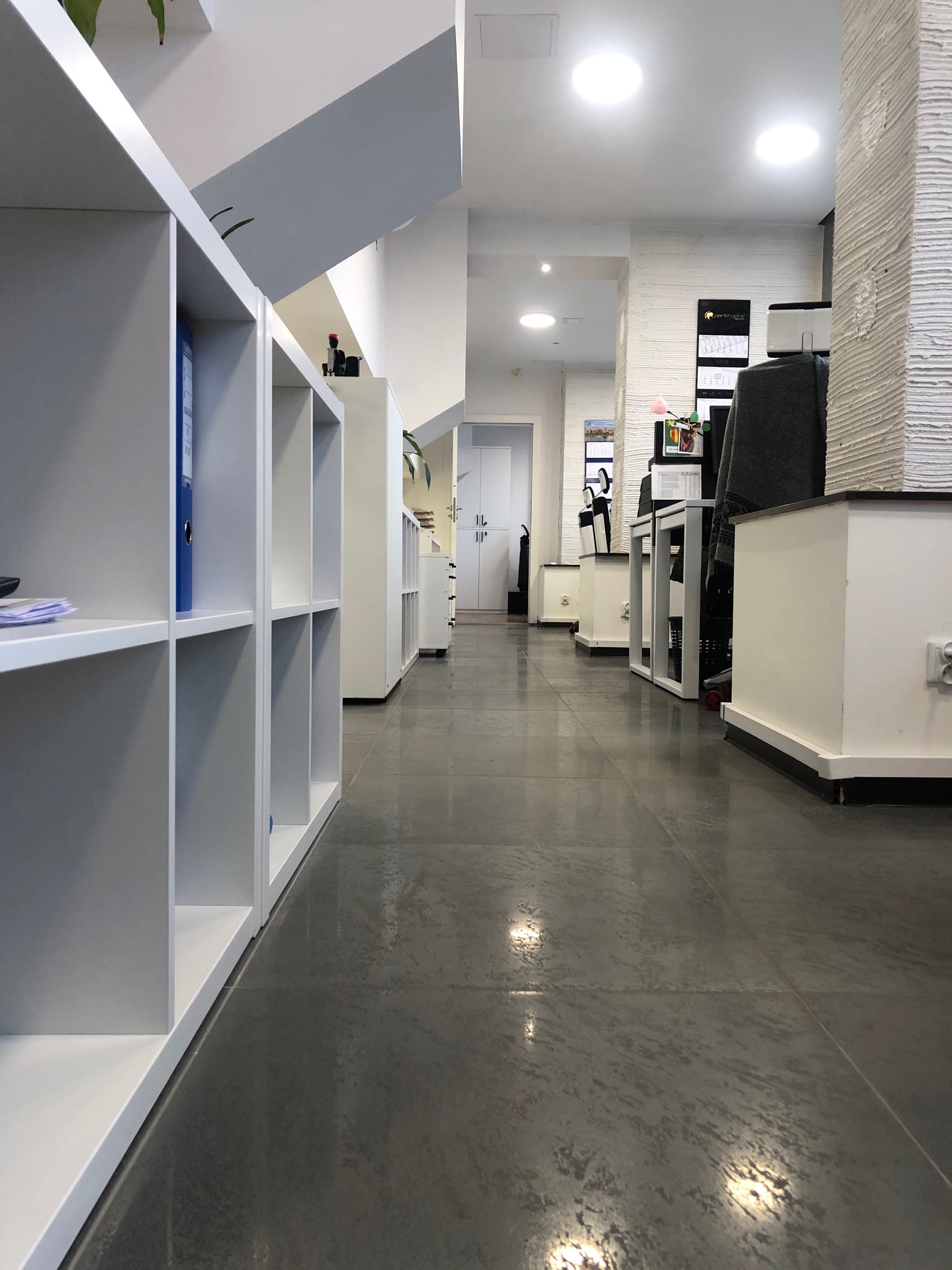 Jasne, przestronne wnętrze biurowe z białymi regałami i szarą podłogą z płytek, widok z perspektywy podłogi wzdłuż korytarza.