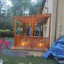 Zadaszony taras z drewnianą konstrukcją, oświetlony lampkami, z widocznym rusztowaniem w tle, częściowo zakryty plandeką.