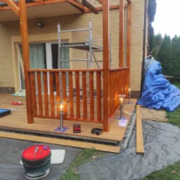 Budowa drewnianej altany z balustradą i oświetleniem zewnętrznym w ogrodzie, widoczne narzędzia, drabina i materiały budowlane na drewnianym podłożu.