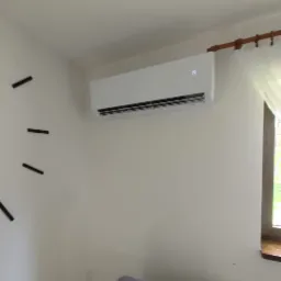 Biała jednostka klimatyzacyjna zamontowana na ścianie w jasnym pokoju, widoczny fragment dekoracyjnego zegara i zasłony okiennej.