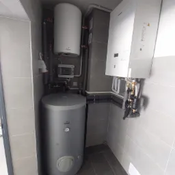 Wnętrze kotłowni z szarymi płytkami, zbiornikiem akumulacyjnym Galmet, wiszącym bojlerem Galmet i kotłem gazowym Panasonic, rury instalacyjne prowadzone wzdłuż ścian.