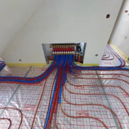 Rozdzielacz ogrzewania podłogowego z czerwonymi i niebieskimi rurami w izolacji, montowany na folii z nadrukiem Roth, widok z góry w surowym stanie.