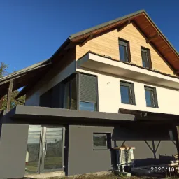 Nowoczesny dom z jasną elewacją i drewnianym wykończeniem poddasza, widok z zewnątrz, na pierwszym planie klimatyzator zewnętrzny i stos drewna.