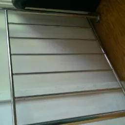 Fragment metalowej balustrady z poziomymi prętami, zamontowanej przy schodach, widoczne materiały reklamowe na ścianie i drewniana podłoga.