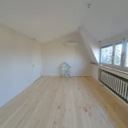 Jasne, przestronne poddasze po remoncie z nową, surową drewnianą podłogą, świeżo pomalowanymi ścianami na biało, grzejnikiem pod oknem i małą białą półką w kształcie domku.