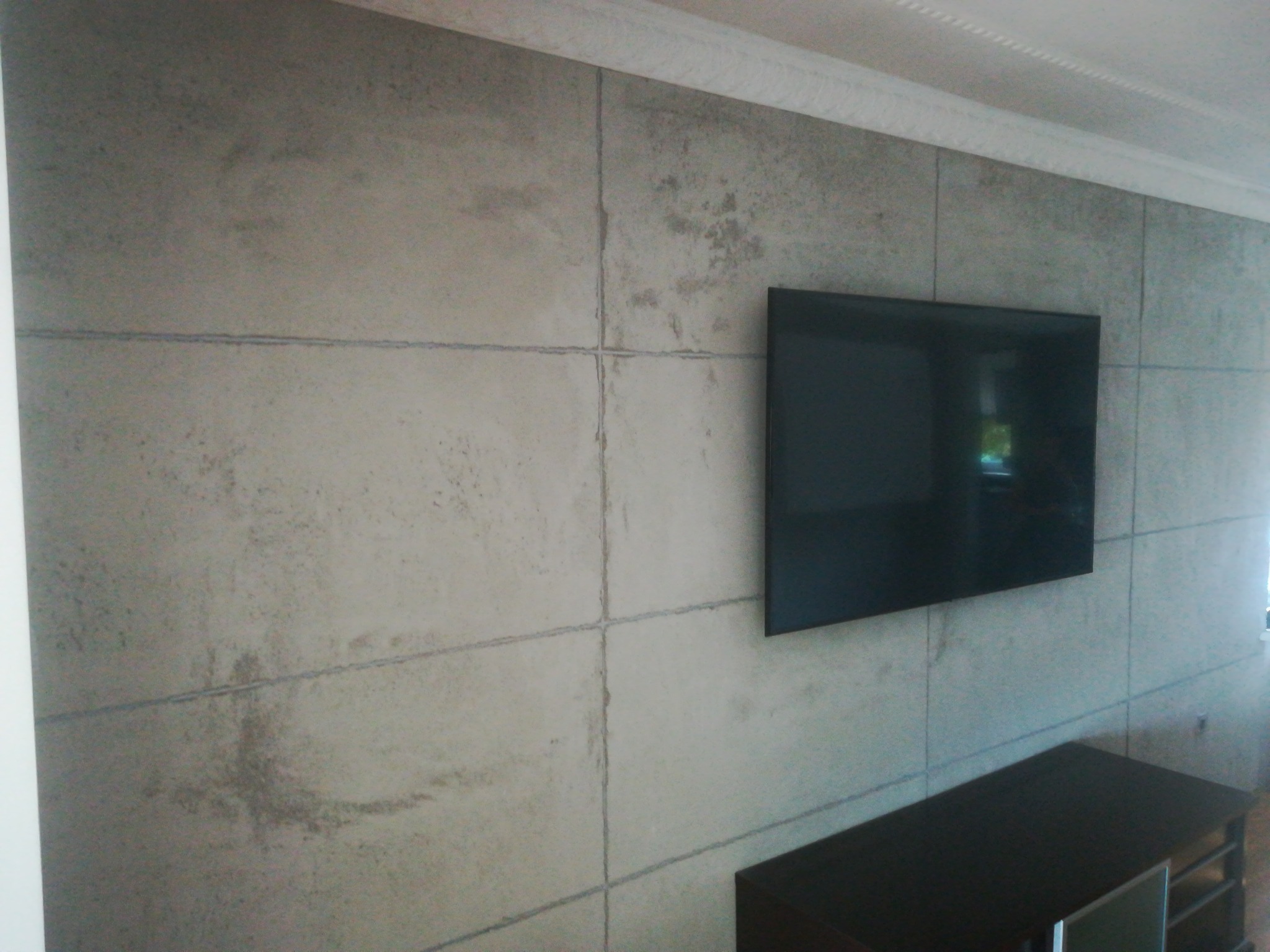 Wykończona ściana z efektem betonu architektonicznego, podzielona na prostokątne panele z widocznymi fugami, z zawieszonym telewizorem i fragmentem ciemnej komody pod nim.