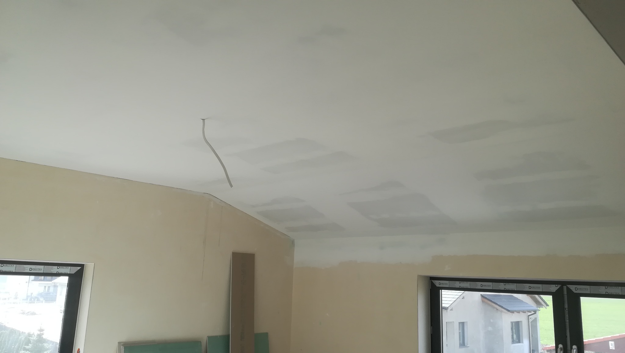 Niedokończone wnętrze z widocznymi łączeniami na suficie, przygotowywanym do dalszych prac wykończeniowych, z wiszącym przewodem elektrycznym i oknami z widokiem na budynek w oddali.